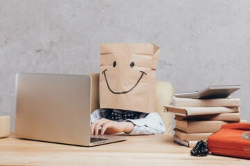 child with brown paper bag on head sitting at laptop