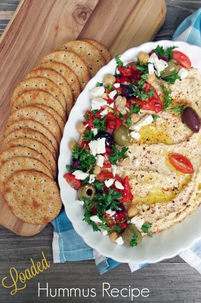 homemade loaded hummus in a white bowl next to a tray of crackers