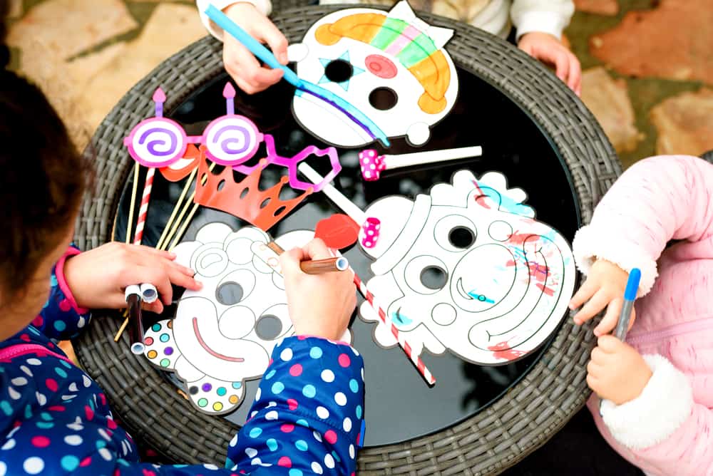 Happy children draw with pencils clown mask. Close up of hands little boy and girls sitting in playroom at the table near Party accessory's and painting. Purim, fools day, Halloween, birthday concept.