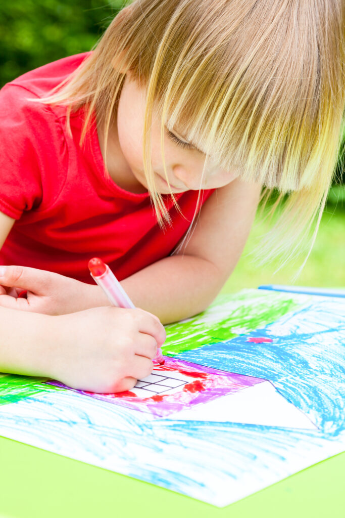 Little girl sitting at table drawing a house outdoors